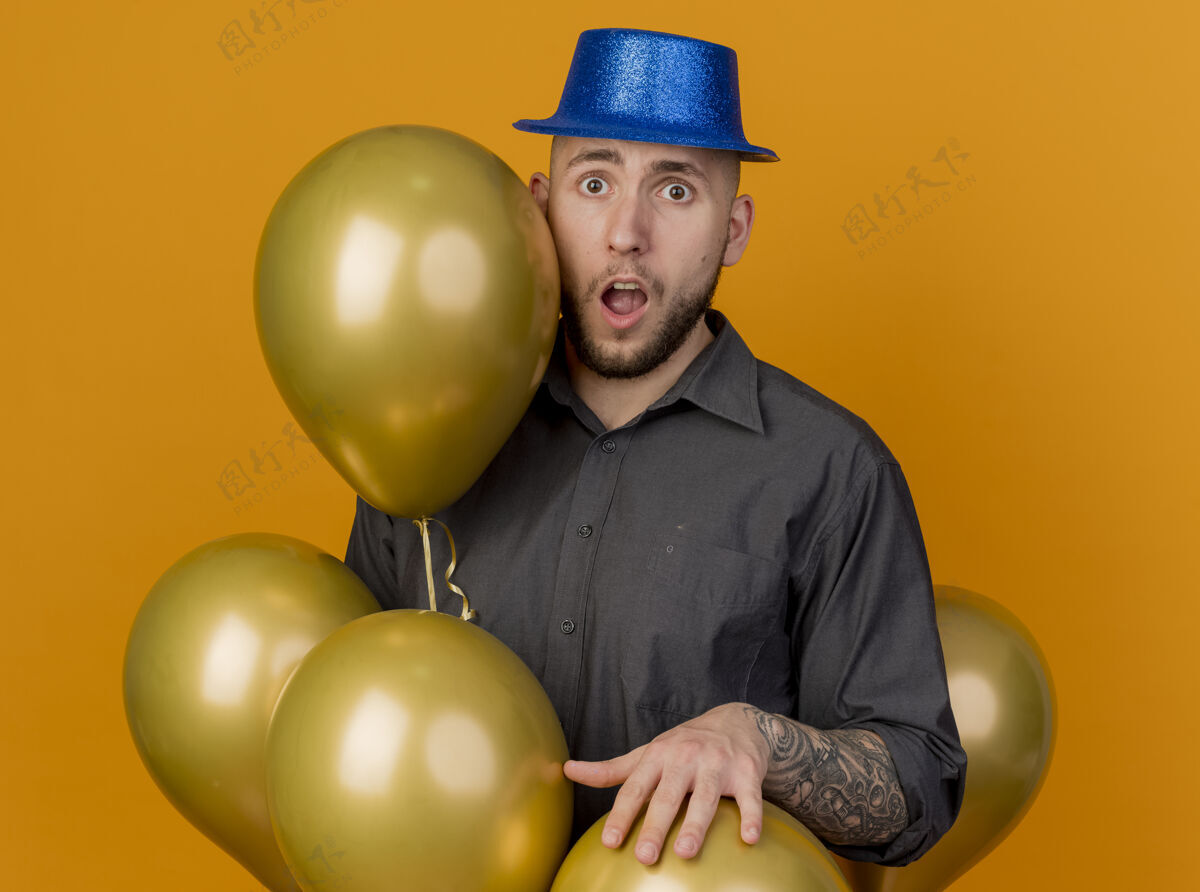 小伙子令人印象深刻的年轻英俊的斯拉夫党的家伙戴着党的帽子站在气球之间把手放在其中一个看着相机孤立的橙色背景印象年轻相机