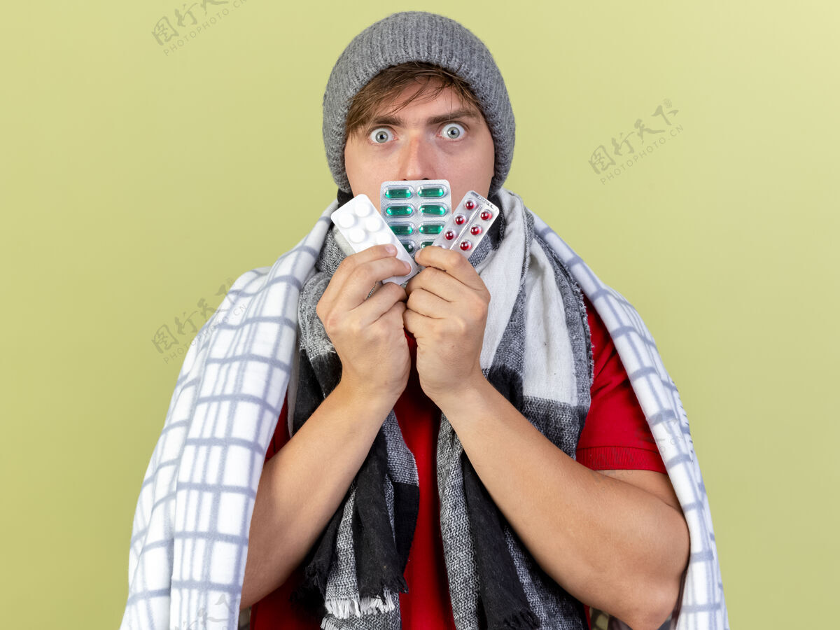 相机令人印象深刻的年轻英俊的金发病男子戴着冬季帽子和围巾包裹在格子布触摸嘴与医疗药丸包看着相机隔离在橄榄绿的背景药片医疗金发