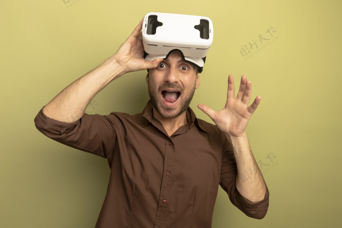 橄榄令人印象深刻的年轻白人男子戴着虚拟现实耳机在前额抓住它看着相机显示空手孤立在橄榄绿的背景与复印空间年轻相机印象