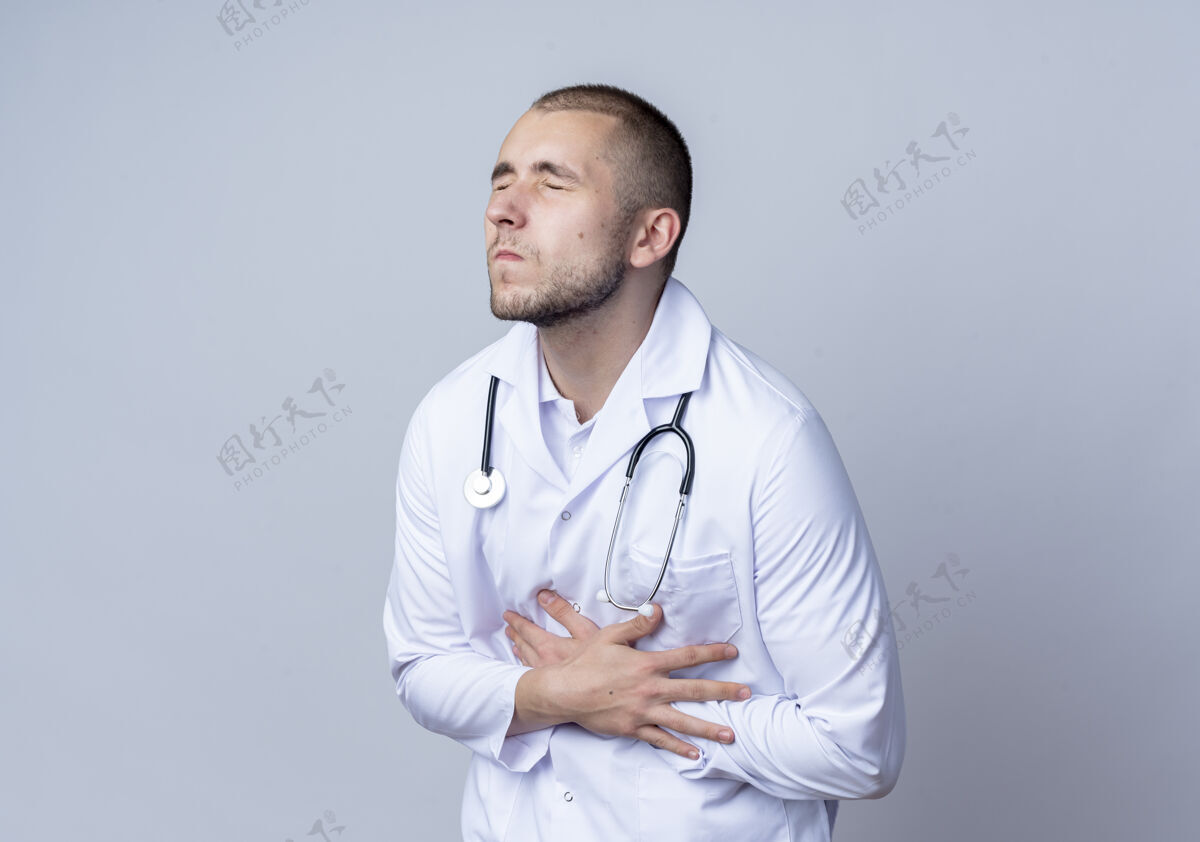眼睛疼痛的年轻男医生 穿着医用长袍 脖子上戴着听诊器 紧闭着眼睛抱着肚子 隔离在白色的空间里疼痛听诊器穿着