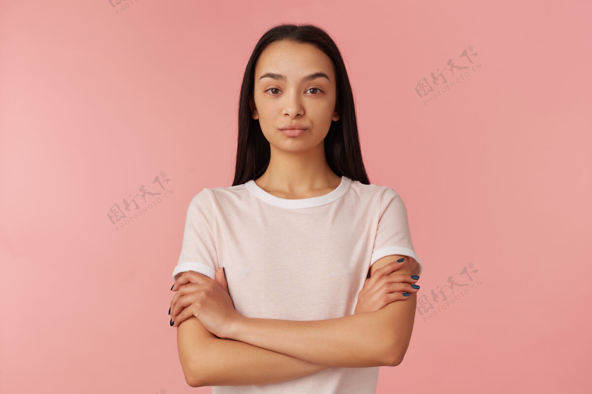 女士一幅严肃 不高兴的女孩的肖像 黑色长发 穿着白色t恤 人和情感的概念 双臂交叉 等待隔离在粉彩粉色的墙上等待交叉表情
