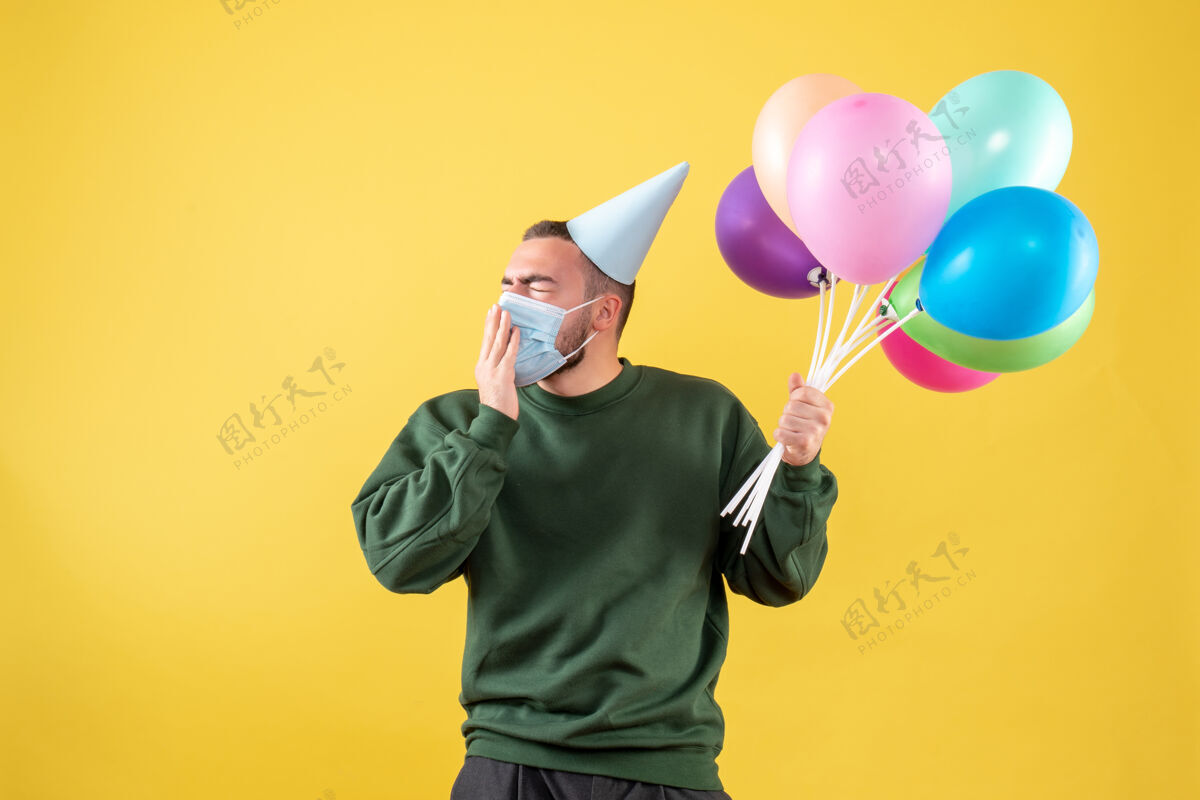 病毒正面图：黄色背景上戴着面具手持彩色气球的年轻男性年轻男性年背景