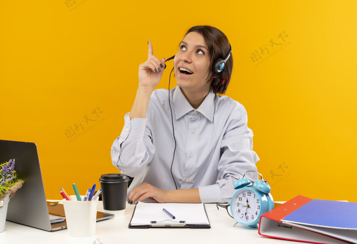 坐着令人印象深刻的年轻的呼叫中心女孩戴着耳机坐在办公桌上看着并指着孤立的橙色电话中心女孩