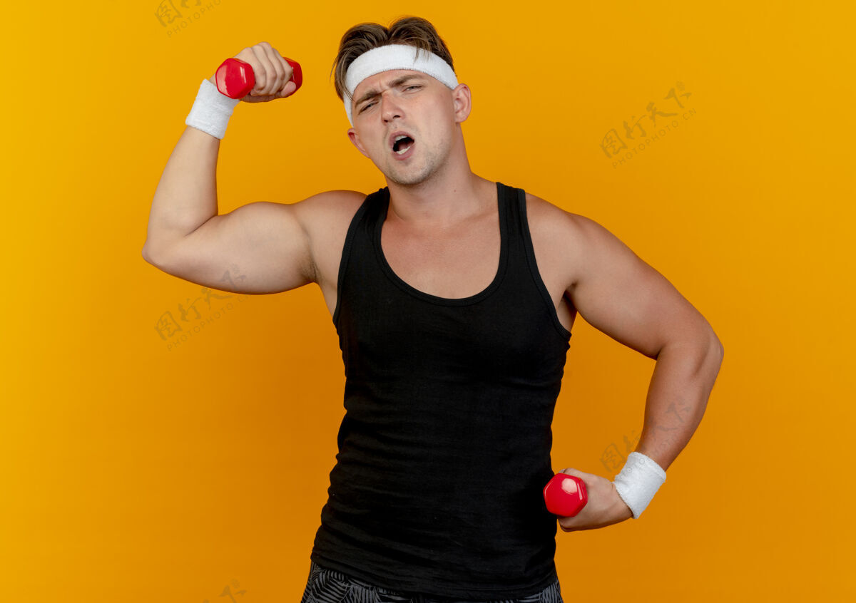 运动自信的年轻英俊的运动型男子戴着头带和腕带举着哑铃孤立在橙色举自信抱着