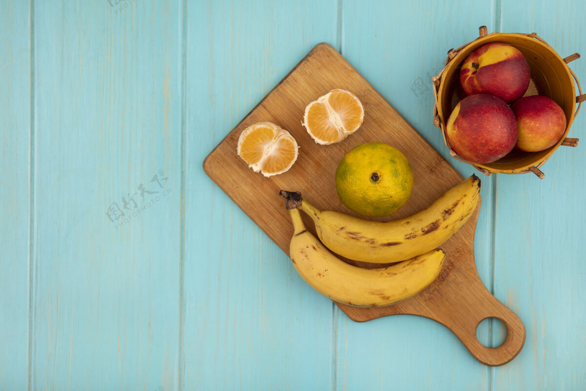 厨房木制厨房板上的整个和半个橘子的俯视图 蓝色木质墙壁上的桶上放着香蕉和桃子 还有复印空间视野树叶香蕉