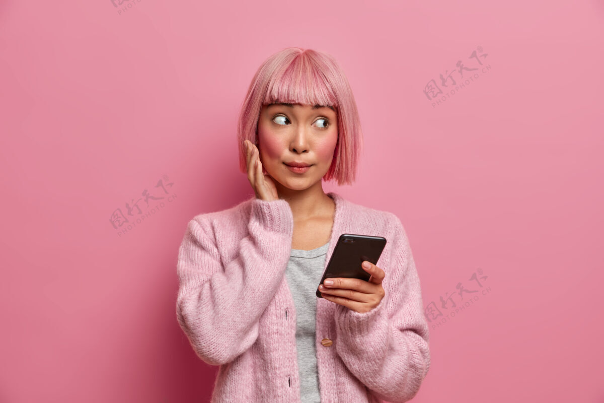 粉色头发一个年轻的亚洲女人脸上露出惊讶的表情 一边看一边 穿着休闲服 拿着手机 在社交媒体上冲浪 在网上发送内容分享多媒体 设备亚洲人姿势