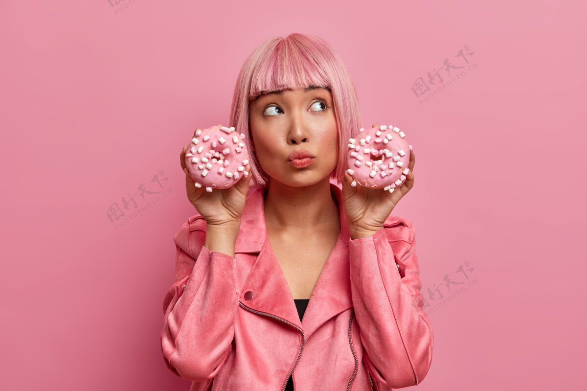 鲍勃梦幻般体贴的亚洲女人拿着两个美味的甜甜圈摆姿势 怀疑是否吃垃圾食品 穿着时髦的粉色夹克 享受甜点人室内粉色假发