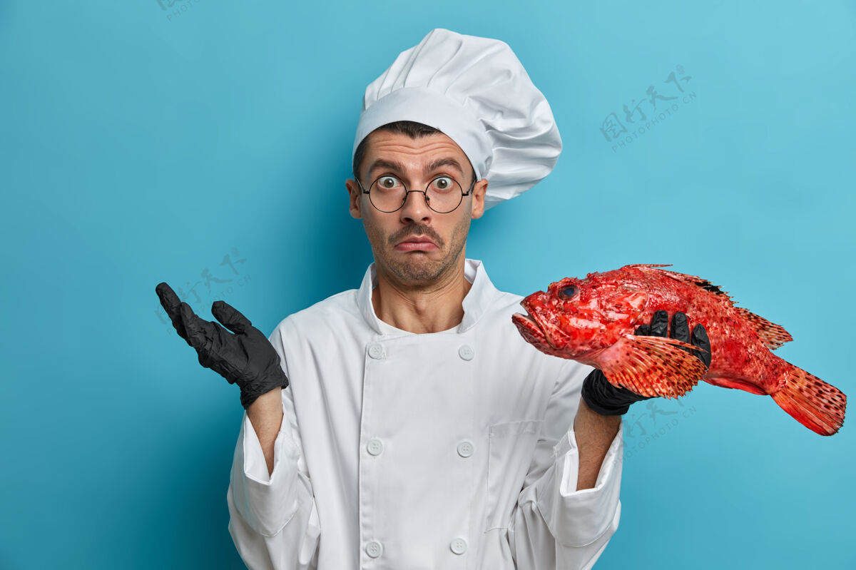手套气愤犹豫不决的厨师手持红海鲈鱼 无法决定要做什么 穿制服 戴黑手套眼镜眼镜烹饪