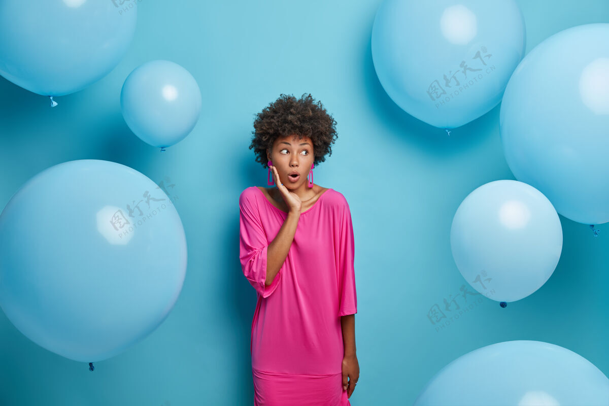 表情惊讶的女人有一头非洲头发 穿着粉红色的节日礼服 震惊地盯着右边 站着聚会庆典风格