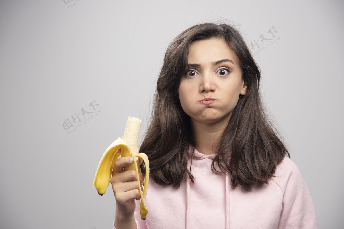 积极年轻的女人在灰色的墙上吃香蕉女孩年轻微笑