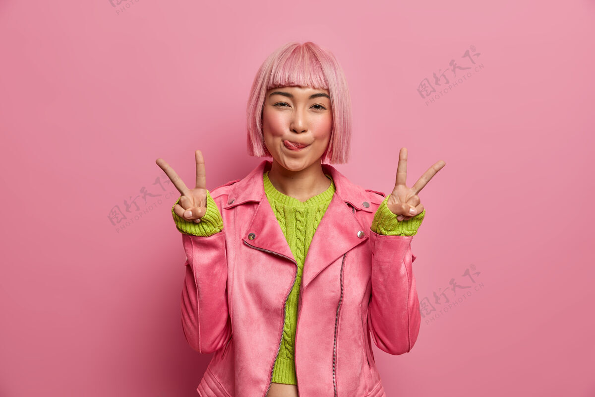 手指积极成功的亚洲粉红头发女人舔嘴唇 做胜利手势 露出两个手指 穿着时尚夹克 在室内摆姿势发型时尚热情