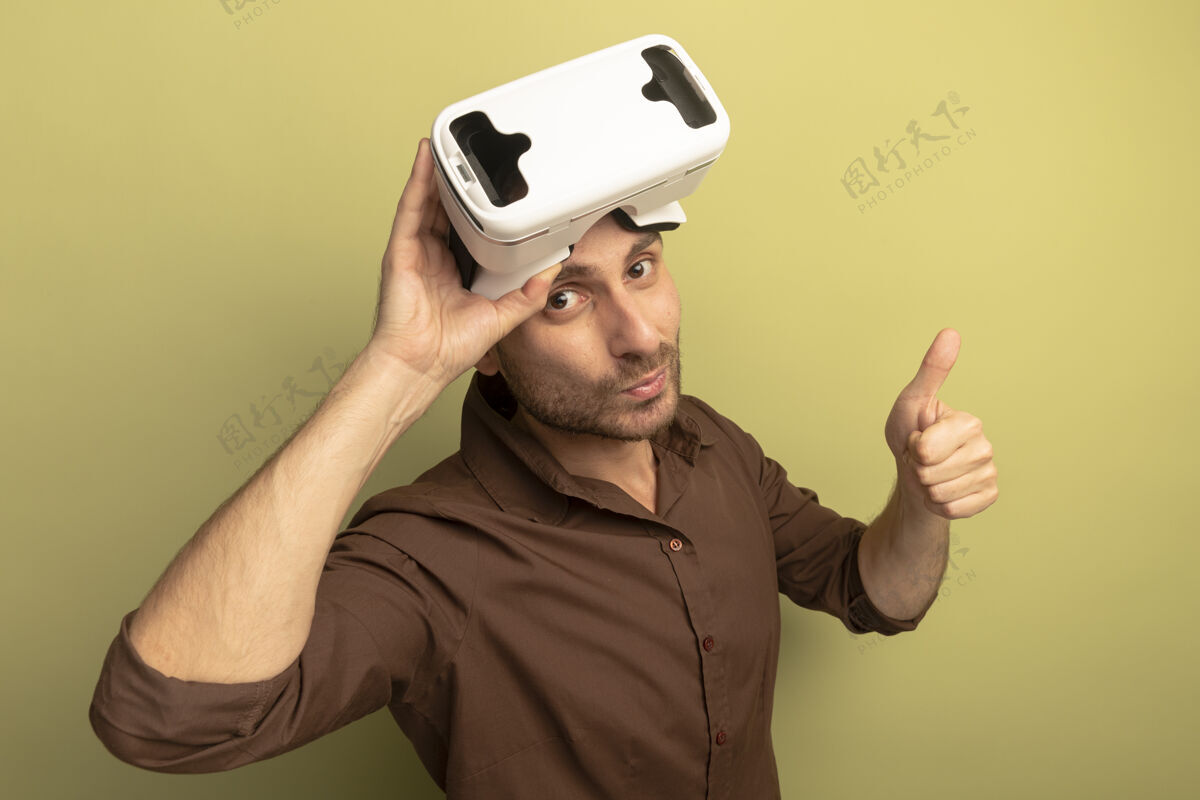 信心自信的白人年轻人头戴虚拟现实耳机 抓住耳机 竖起大拇指 看着橄榄绿背景上的相机穿展示男人