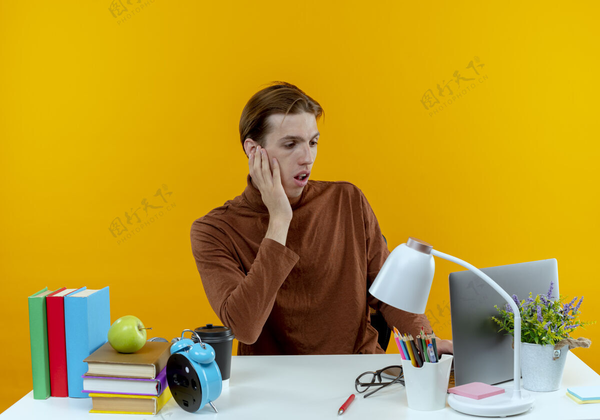 笔记本电脑惊讶的年轻学生男孩坐在书桌旁 手里拿着笔记本电脑用的学习工具 把手放在黄色的脸颊上学校黄色年轻