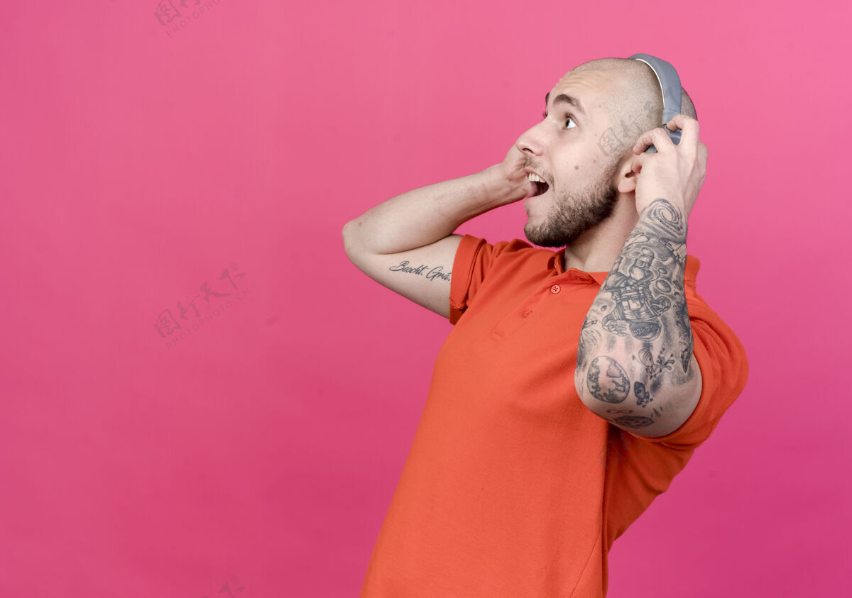 侧线看着旁边惊讶的年轻运动男子抓住耳机在他的头上隔离在粉红色的墙壁与复制空间墙耳机头