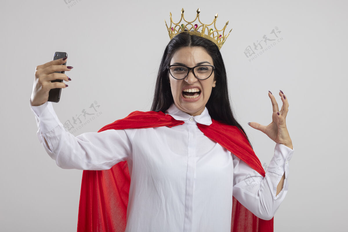 感情愤怒的年轻女超人戴着眼镜 戴着皇冠 手里拿着手机 手举在空中看着隔离在白墙上的前方人穿着人