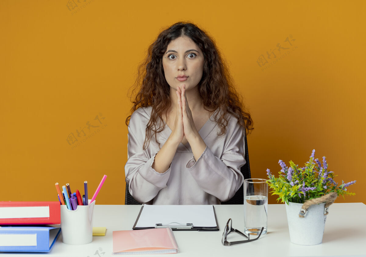 年轻人一位年轻漂亮的女上班族坐在办公桌旁 手里拿着办公工具 在橙色的桌子上显示出祈祷的姿势办公桌工具女性