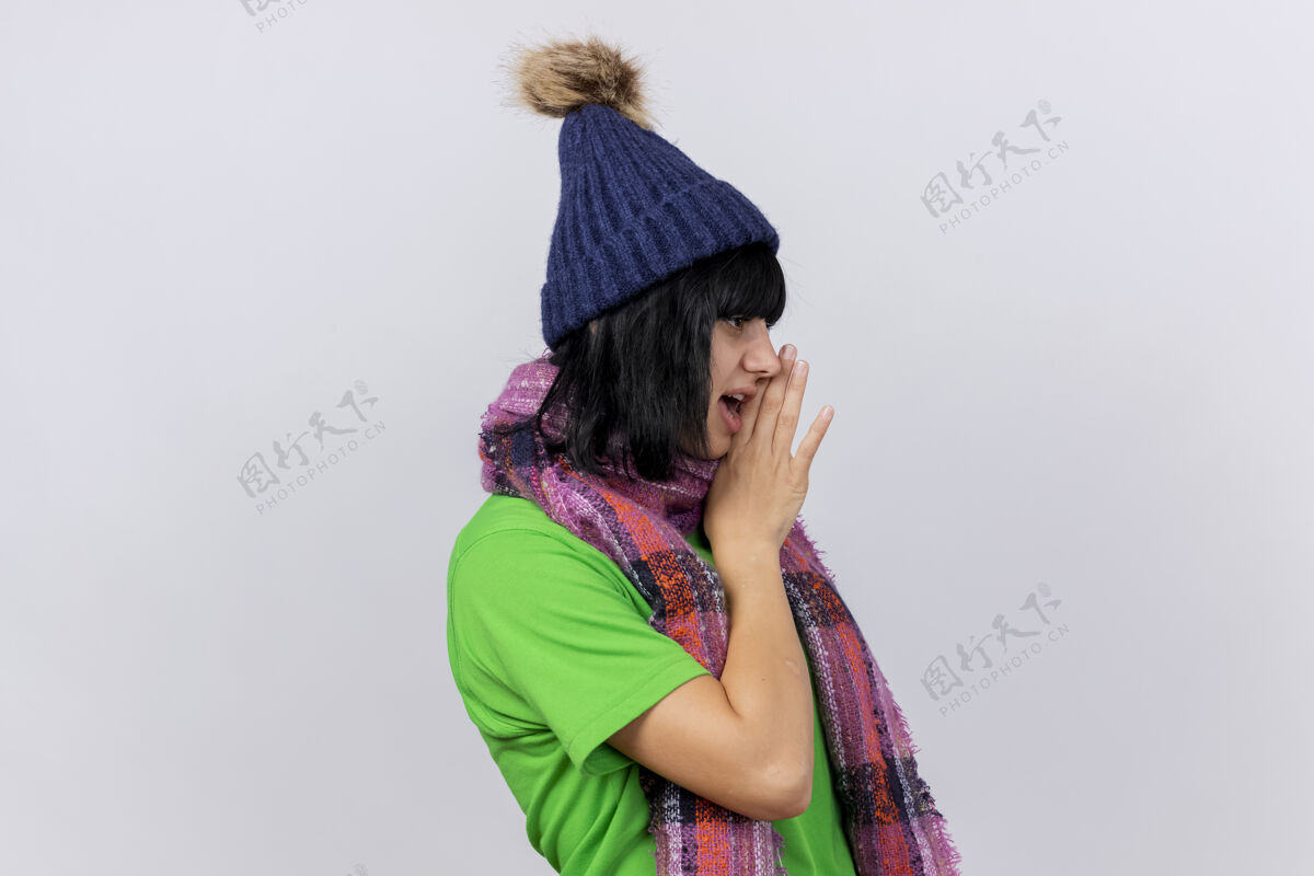 耳语戴着冬日帽和围巾的年轻病妇站在侧视图中看着笔直的手靠近嘴在白色的墙上低语着隔离表情手嘴