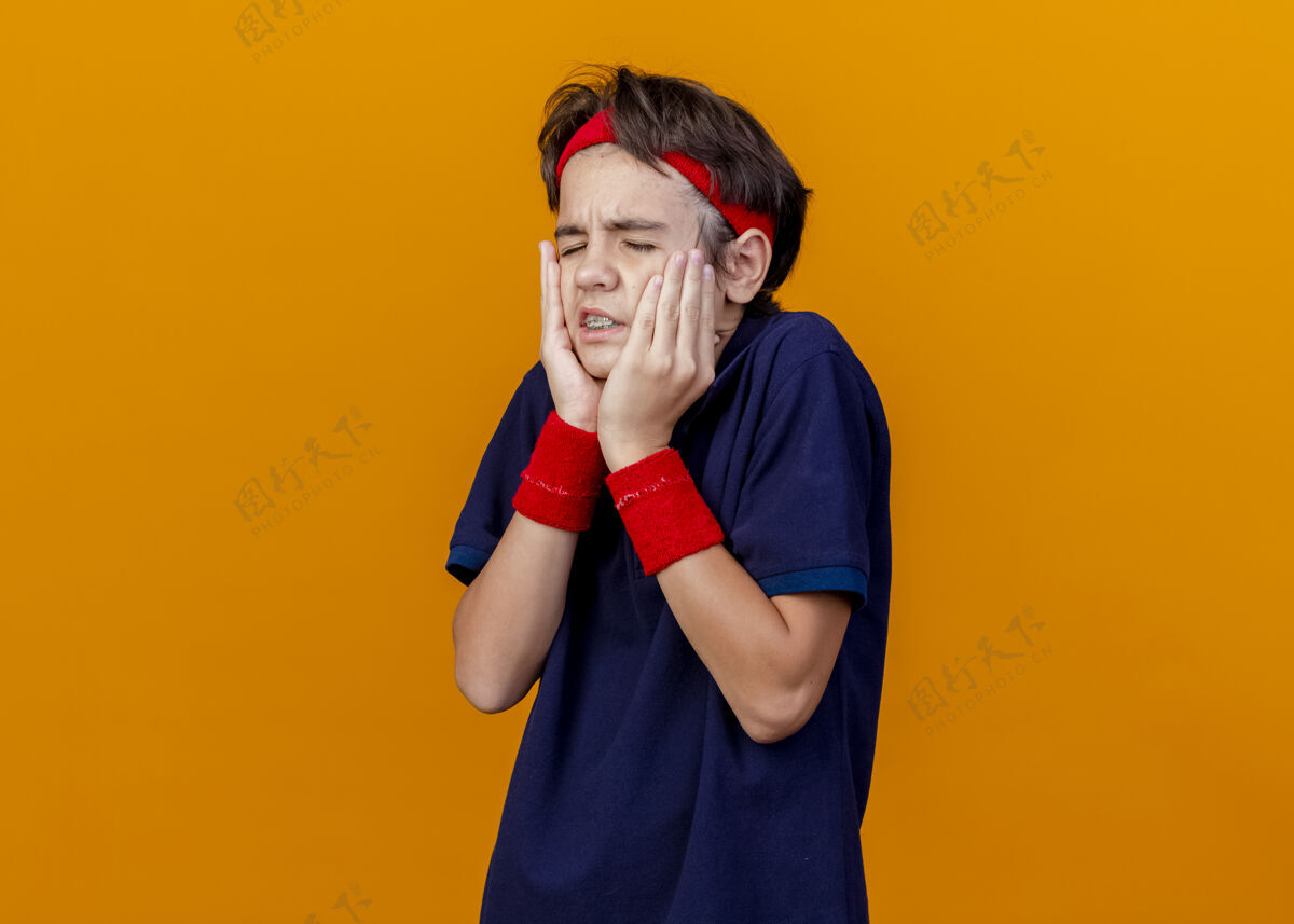人疼痛的年轻英俊的运动男孩戴着头带和戴着牙套的腕带 手放在脸上 闭着眼睛孤立在橙色的墙上脸手穿着