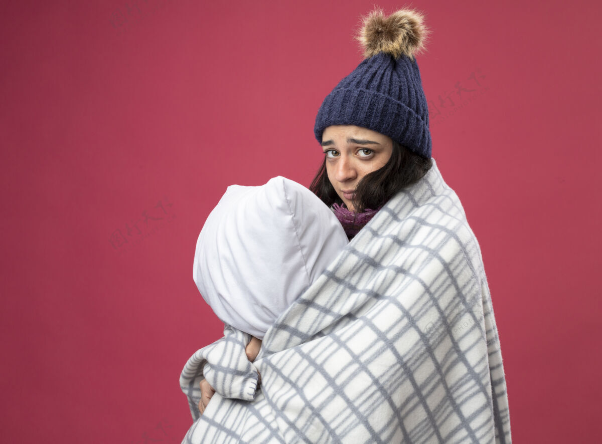 疾病体弱多病的年轻女子穿着长袍 戴着冬天的帽子 围着方格呢围巾站在侧视图里 抱着枕头 看着隔离在粉色墙上的前面脸感觉轮廓
