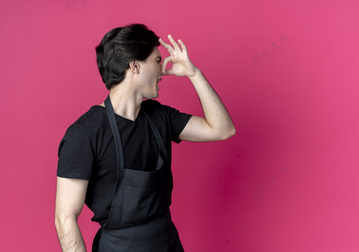 侧面站在侧视图上年轻帅气的男理发师穿着制服闭着鼻子孤立地站在粉色带拷贝的空间里男性空间制服
