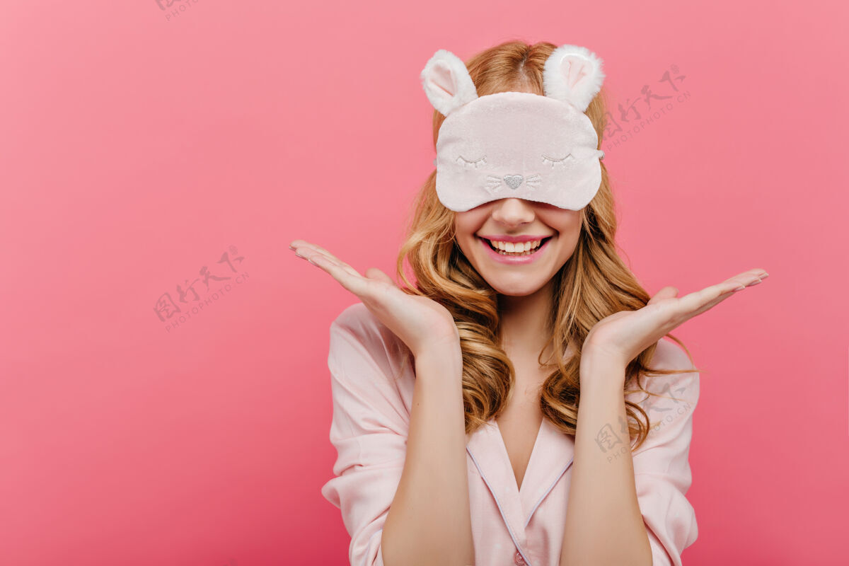 女人戴着眼罩的快乐美丽的年轻女子穿着睡衣的快乐的欧洲女孩早上戴着睡眠面罩站在粉红色的墙上年轻表情睡衣