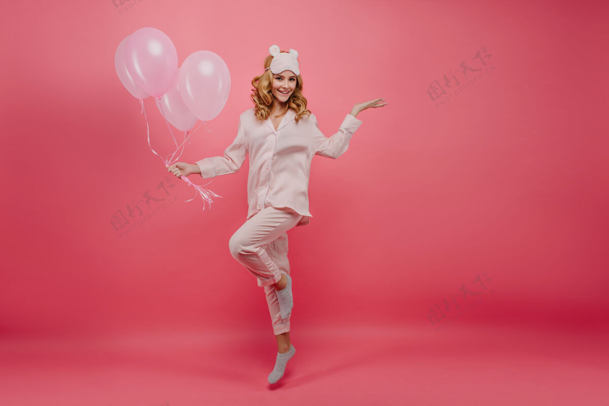氦气球穿灰色袜子的生日女孩摆姿势的全长照片穿着丝绸睡衣的微笑的年轻女士和粉色气球跳跃的肖像美丽节日舞蹈