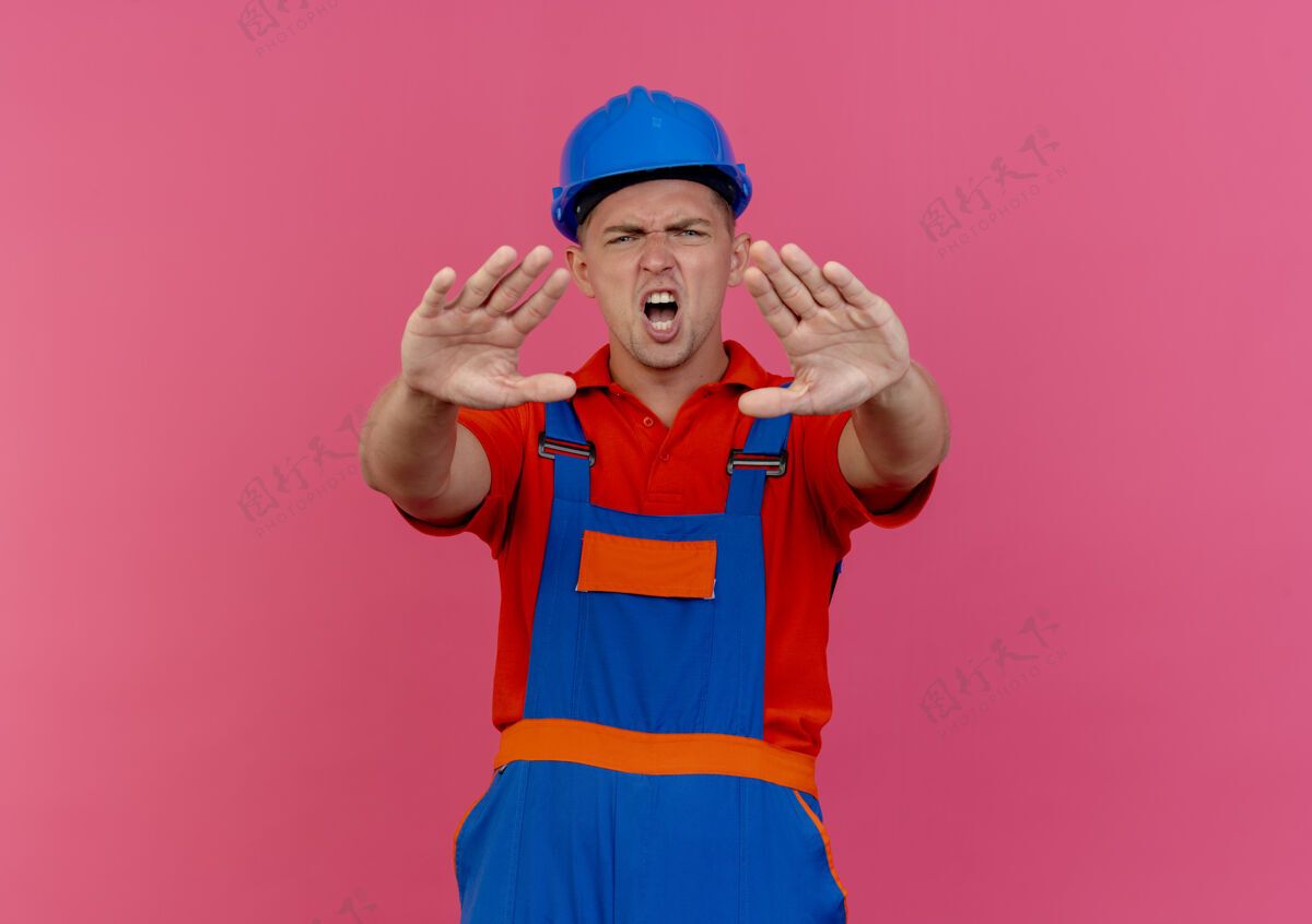 头盔未出租的年轻男性建筑工人穿着制服 戴着安全帽 在粉红色的地板上做停车手势建筑工人制服手势