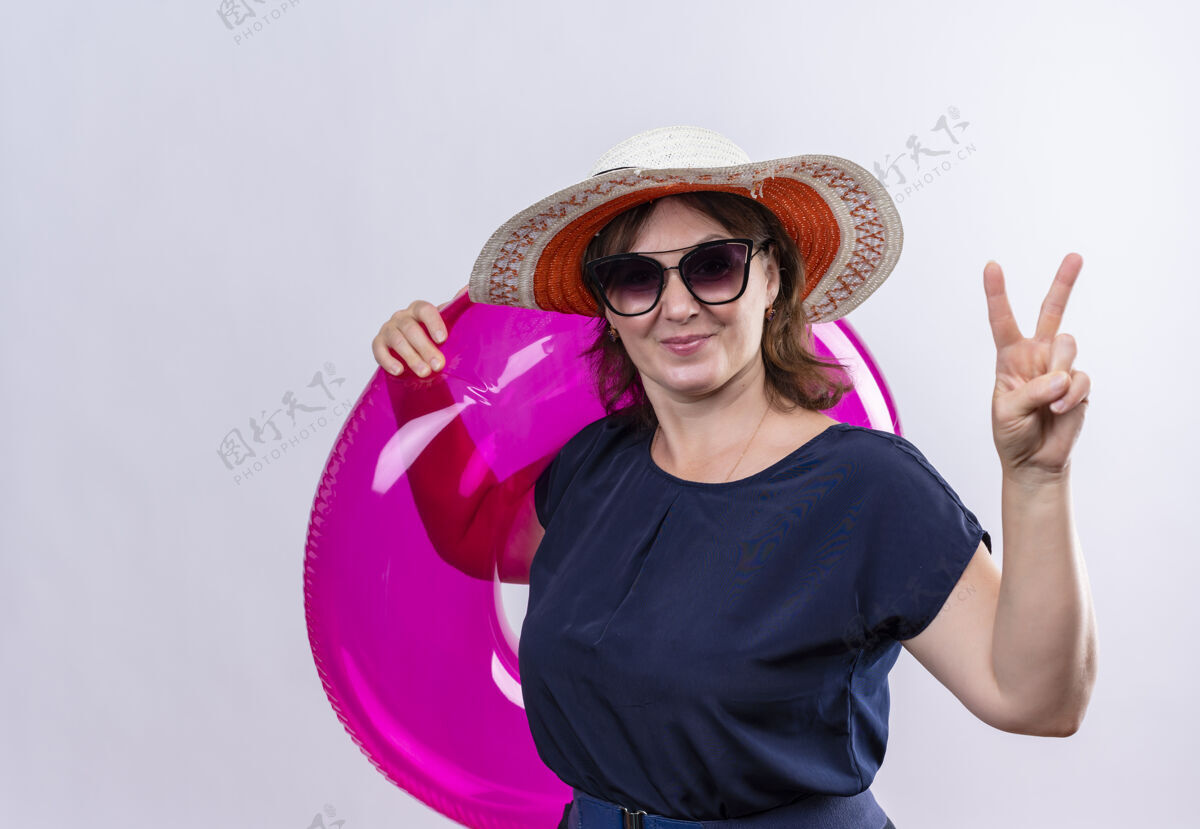 穿一位戴着眼镜 戴着帽子 手持充气戒指的中年旅行家女士在与世隔绝的白墙上展示着和平的姿态展示中年充气