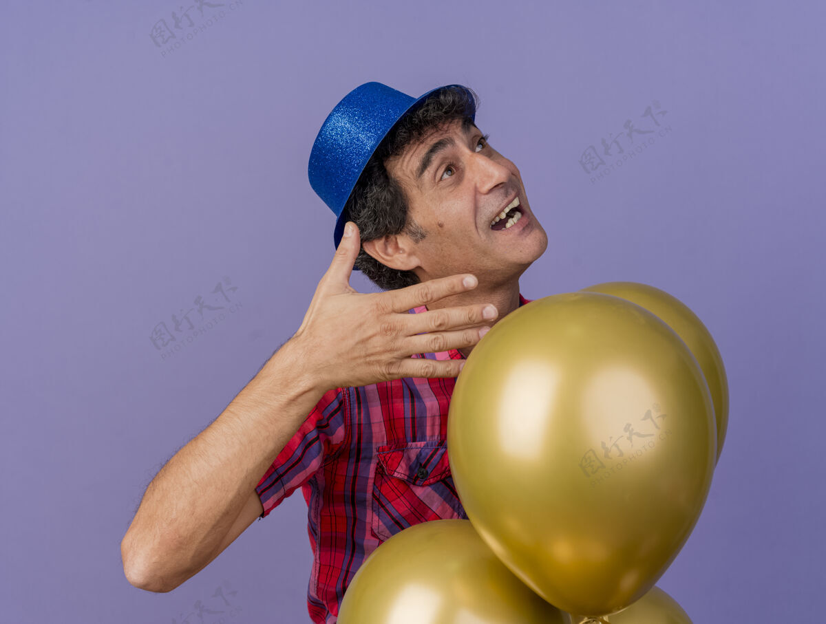 空气令人印象深刻的中年党人戴着党的帽子站在气球后面保持手在空中看着紫色墙上孤立穿着帽子人