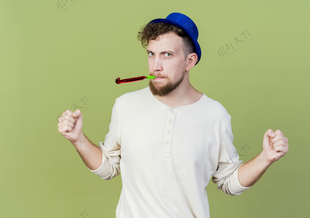 男人愁眉苦脸的年轻帅哥戴着派对帽 紧握拳头 看着橄榄绿墙上的吹风机皱眉站穿