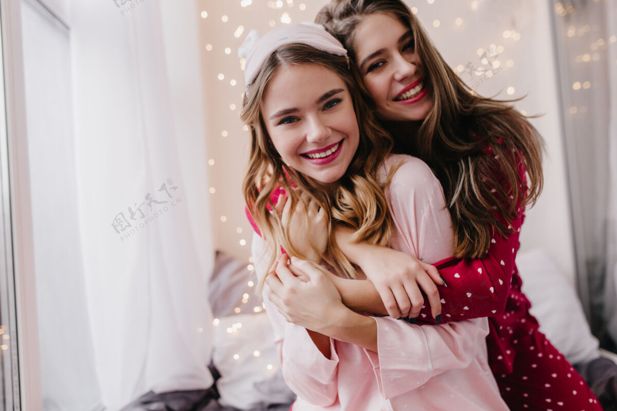 室内室内拍摄的幸福卷发女孩摆出真诚的微笑在她舒适的房间里优雅的欧洲女模特拥抱妹妹和笑黑发快乐享受