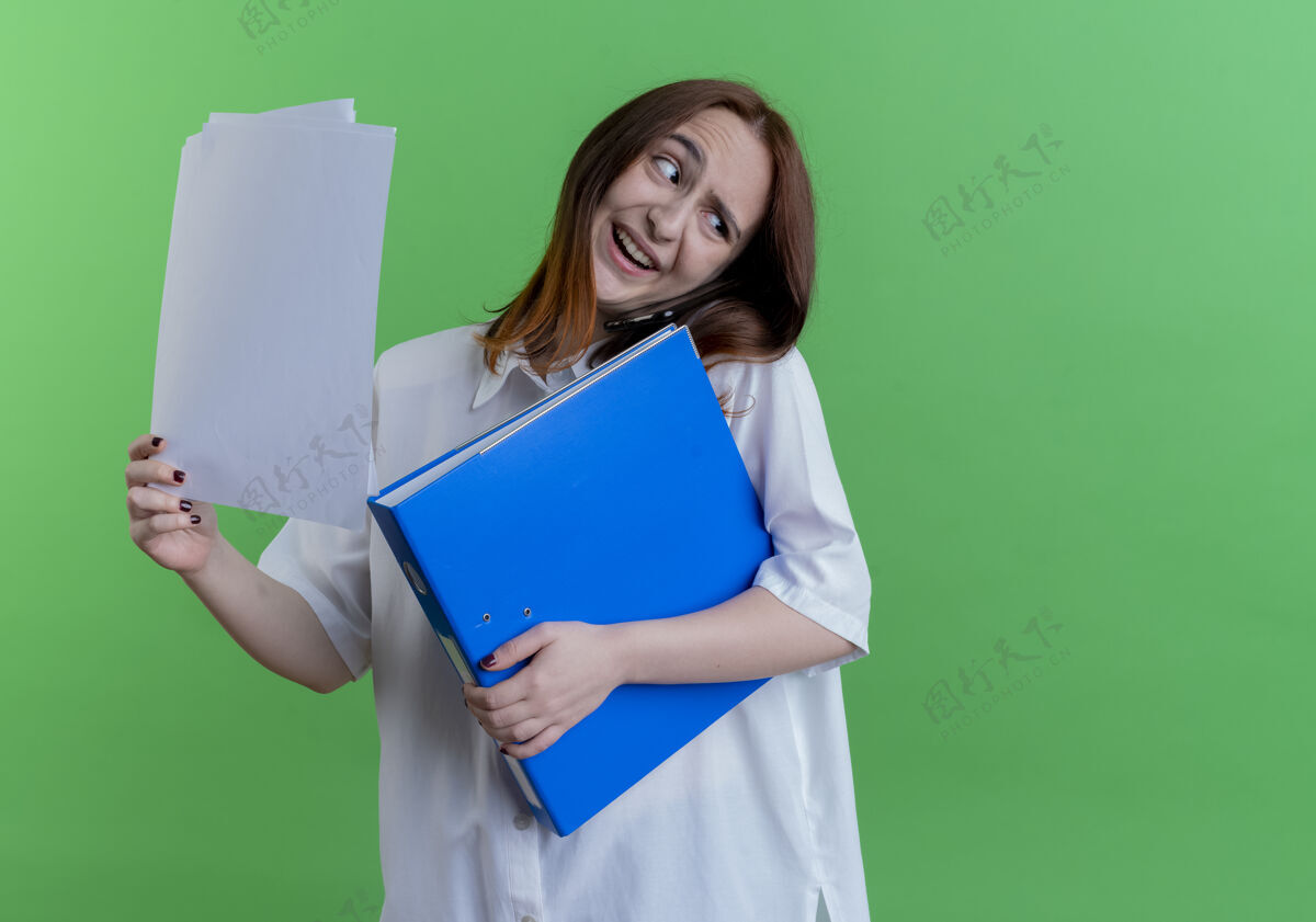 说话歪着头微笑的年轻红发女孩拿着文件夹纸 在绿色的隔离地上讲电话纸握着文件夹