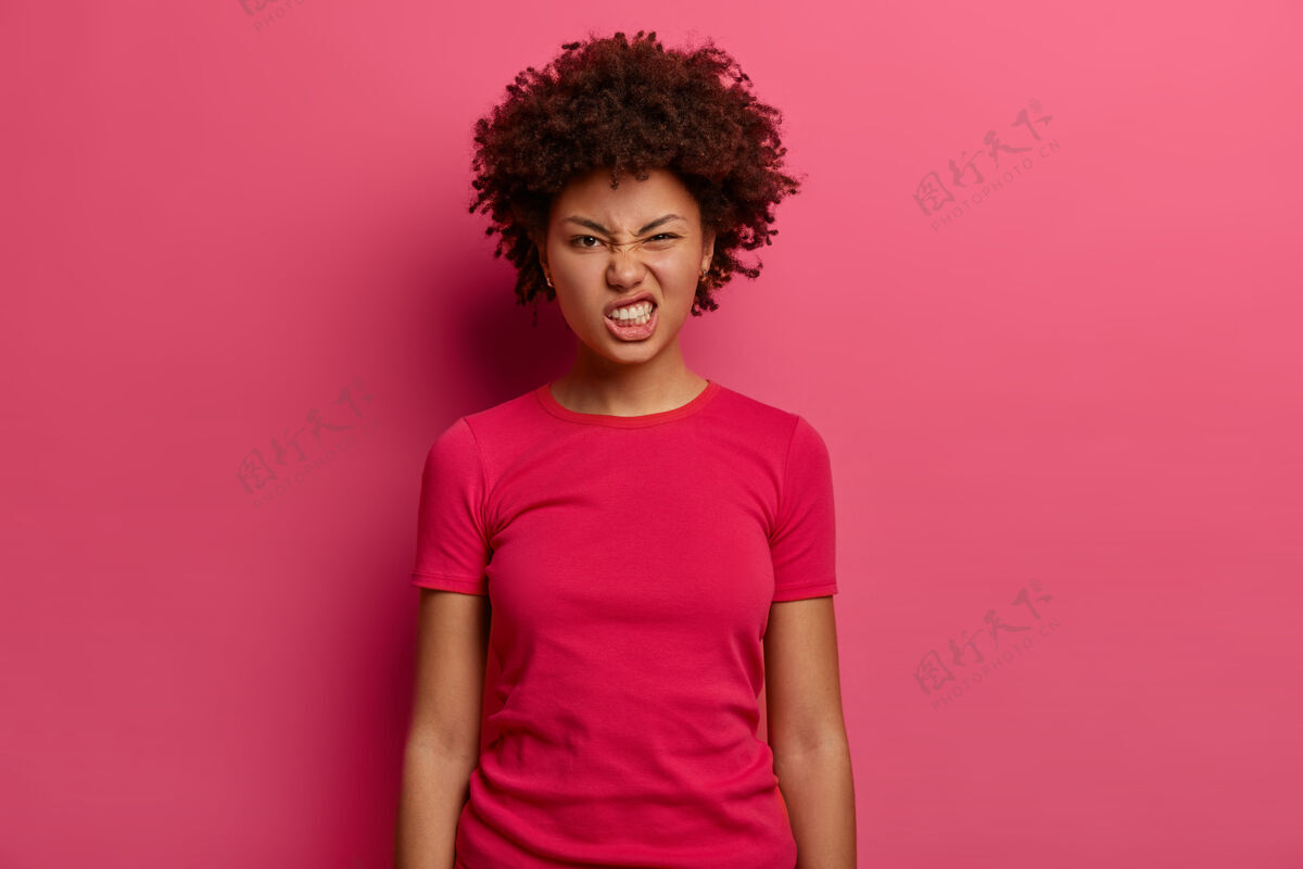 厌恶恼怒不满意的女人咬牙切齿感到恼怒 看起来很不高兴 穿着休闲t恤 在室内对着粉色墙壁摆姿势消极的面部表情概念女孩单色面部