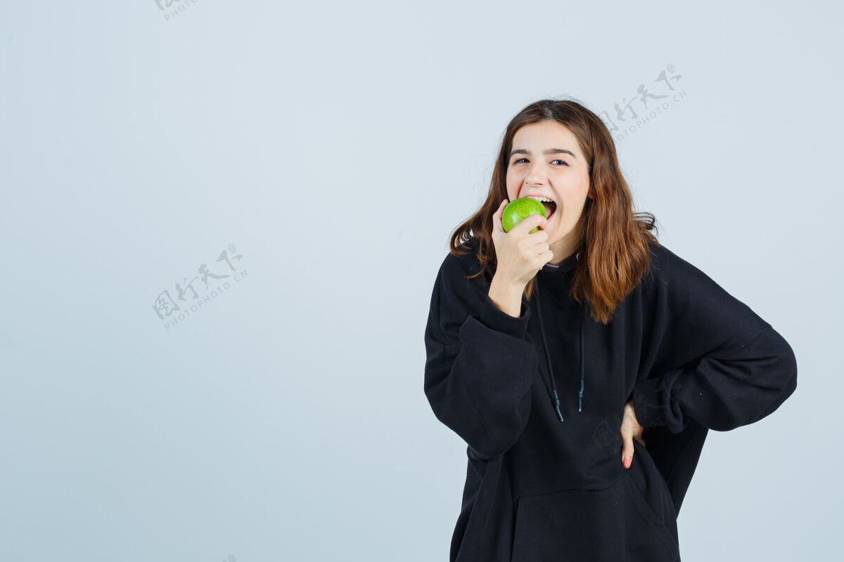 腰年轻女士咬着苹果 手放在腰上 穿着超大的连帽衫和裤子 看起来很高兴正视图护理头发健康