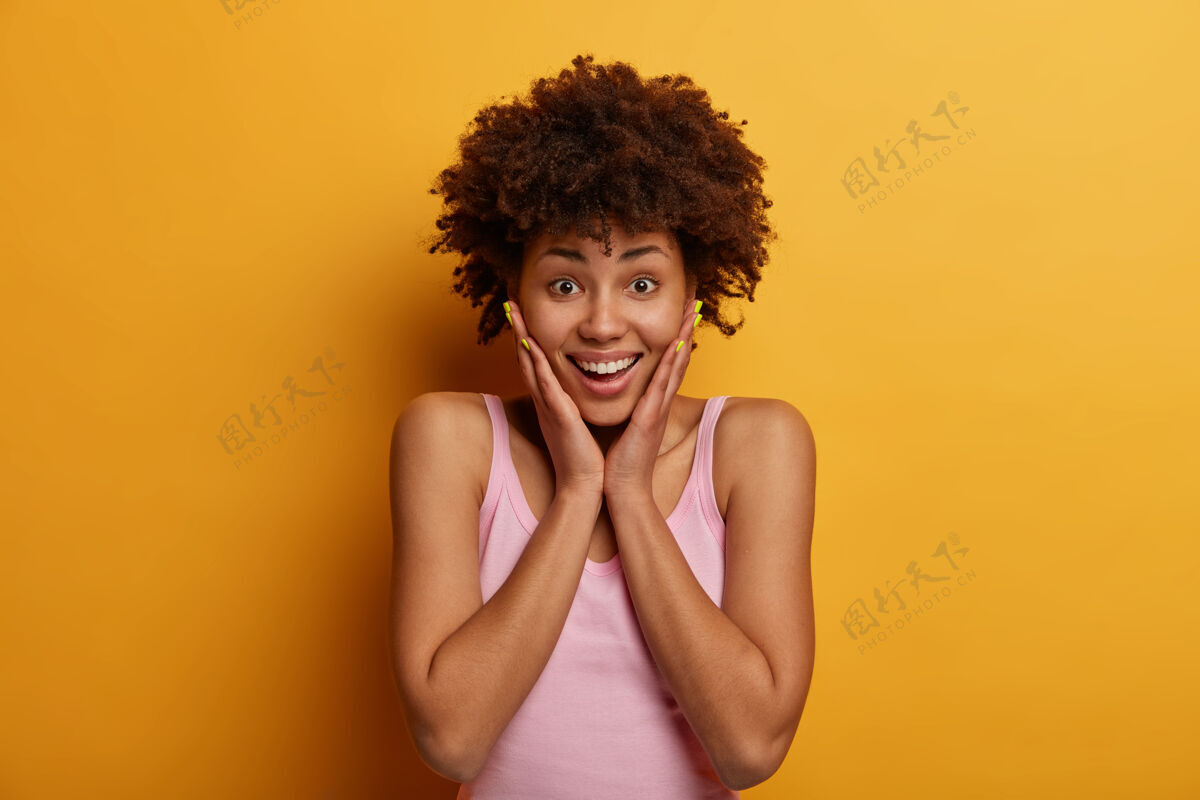 真诚积极的情绪概念快乐快乐的卷曲的非洲裔美国妇女触摸脸颊 学到了一些意想不到的和可怕的东西 带着快乐的微笑看着 在黄色的墙上摆姿势脸高兴有趣