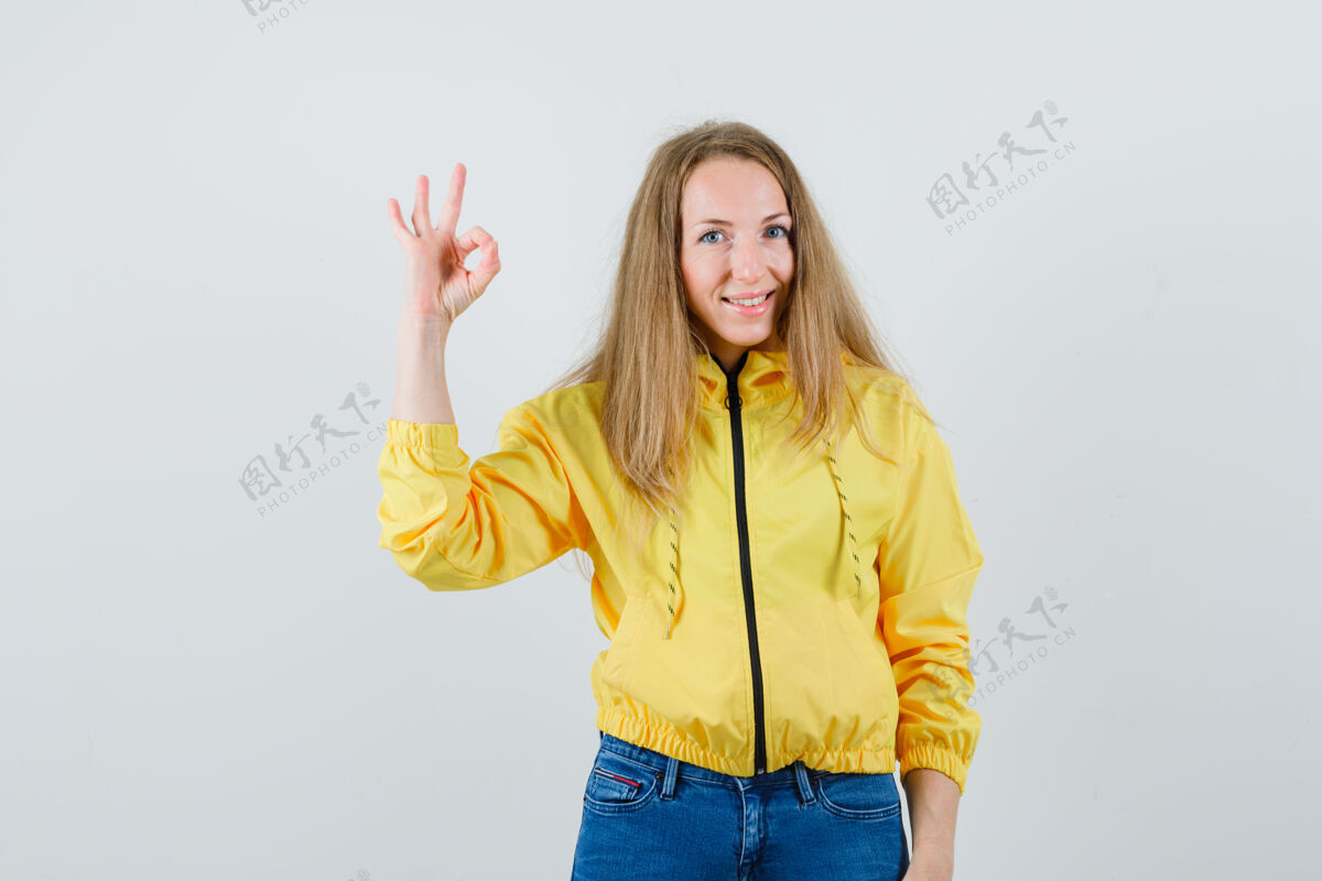 肖像身穿黄色棒球服和蓝色牛仔裤 表情严肃的年轻女子 展示着“OK”的手势和沉默的手势好的年轻时髦