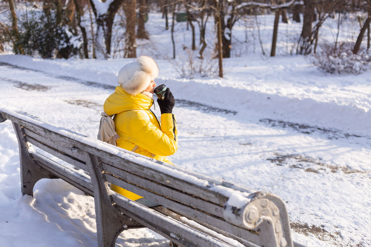 咖啡冬天的快乐年轻女子 穿着暖和的衣服 在阳光明媚的日子里 坐在长椅上 独自享受着新鲜的空气和咖啡快乐冬天的衣服杯子