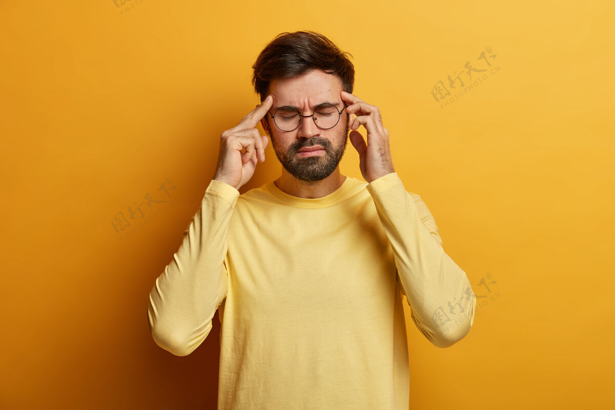 太阳穴过度劳累沮丧的胡须男子按摩太阳穴 患有严重的偏头痛 闭上眼睛缓解疼痛 戴着眼镜和休闲的黄色毛衣 站在那里 试图冷静下来眼镜傻笑头痛