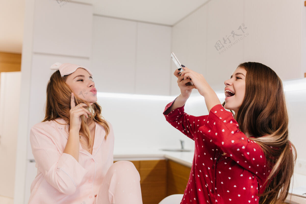 睡衣迷人的卷发女孩摆出亲吻的表情笑着穿红衬衫的欧洲女人用手机为她可爱的妹妹拍照成人电话睡衣