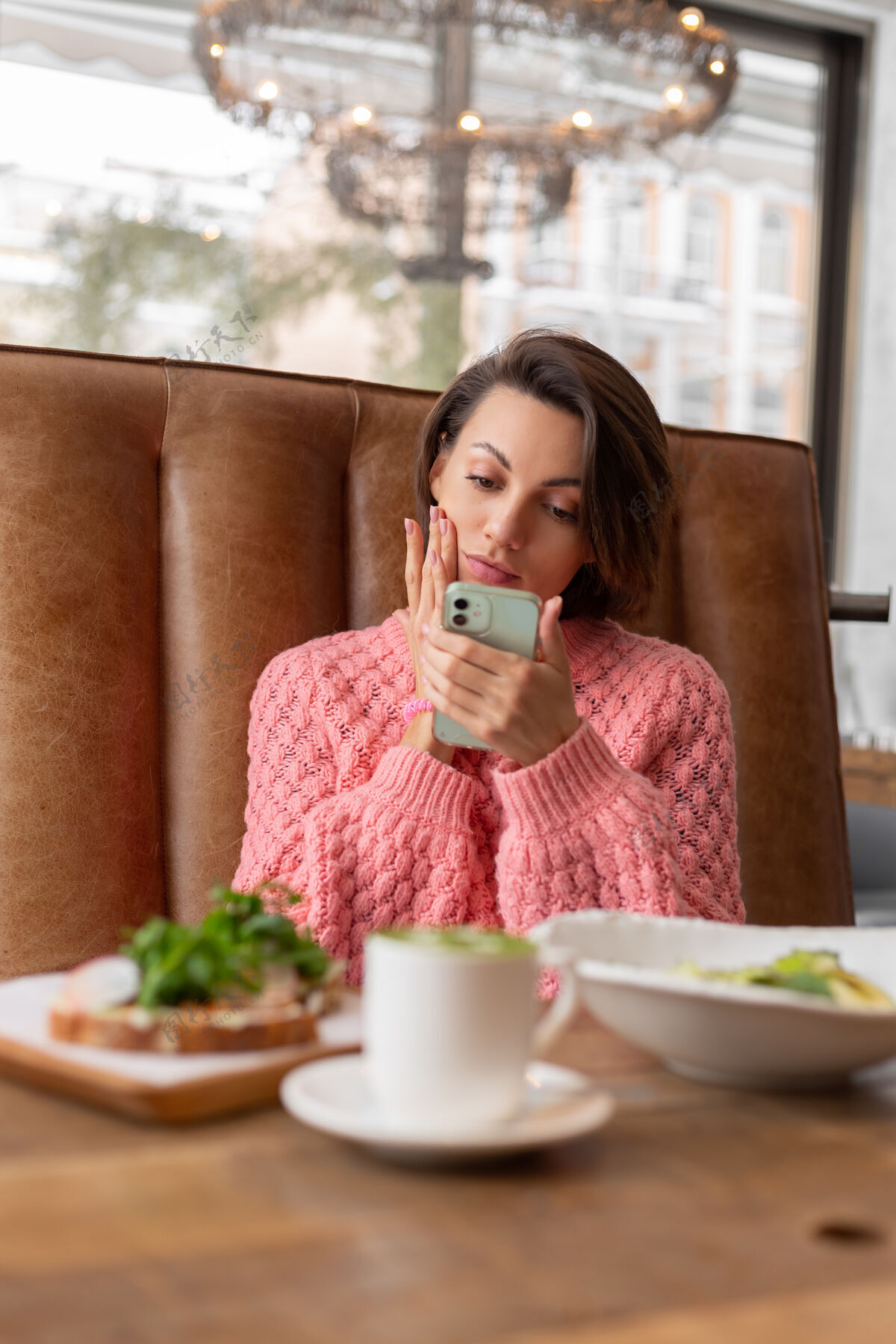 美味餐厅里一位穿着暖和毛衣的女士正在看电话里的东西 桌上摆着美味健康的早餐和抹茶拿铁芝麻菜切片电话