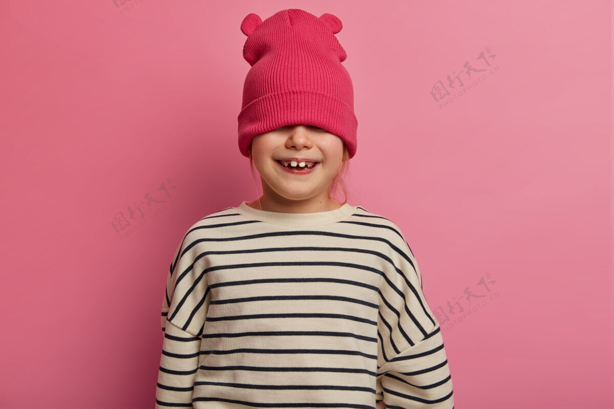 红发顽皮的小女孩戴着时髦的帽子 藏着眼睛 玩得很开心 不想去幼儿园 有着牙齿般的微笑 心情非常乐观 在柔和的玫瑰色墙壁上摆姿势孩子 时尚 时尚隐藏姜罗西