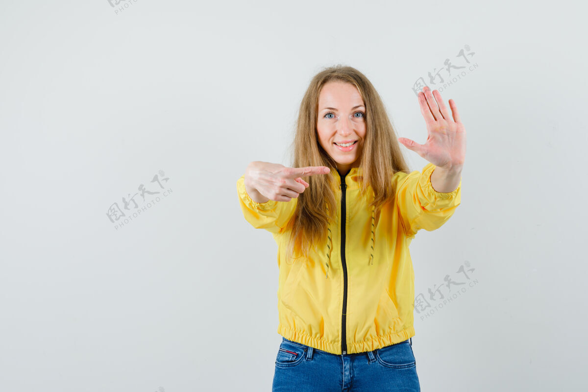 指向一只手举着停车标志的年轻女子 穿着黄色棒球服和蓝色牛仔裤 指着标志 看上去很乐观 正对着前方年轻时尚女人