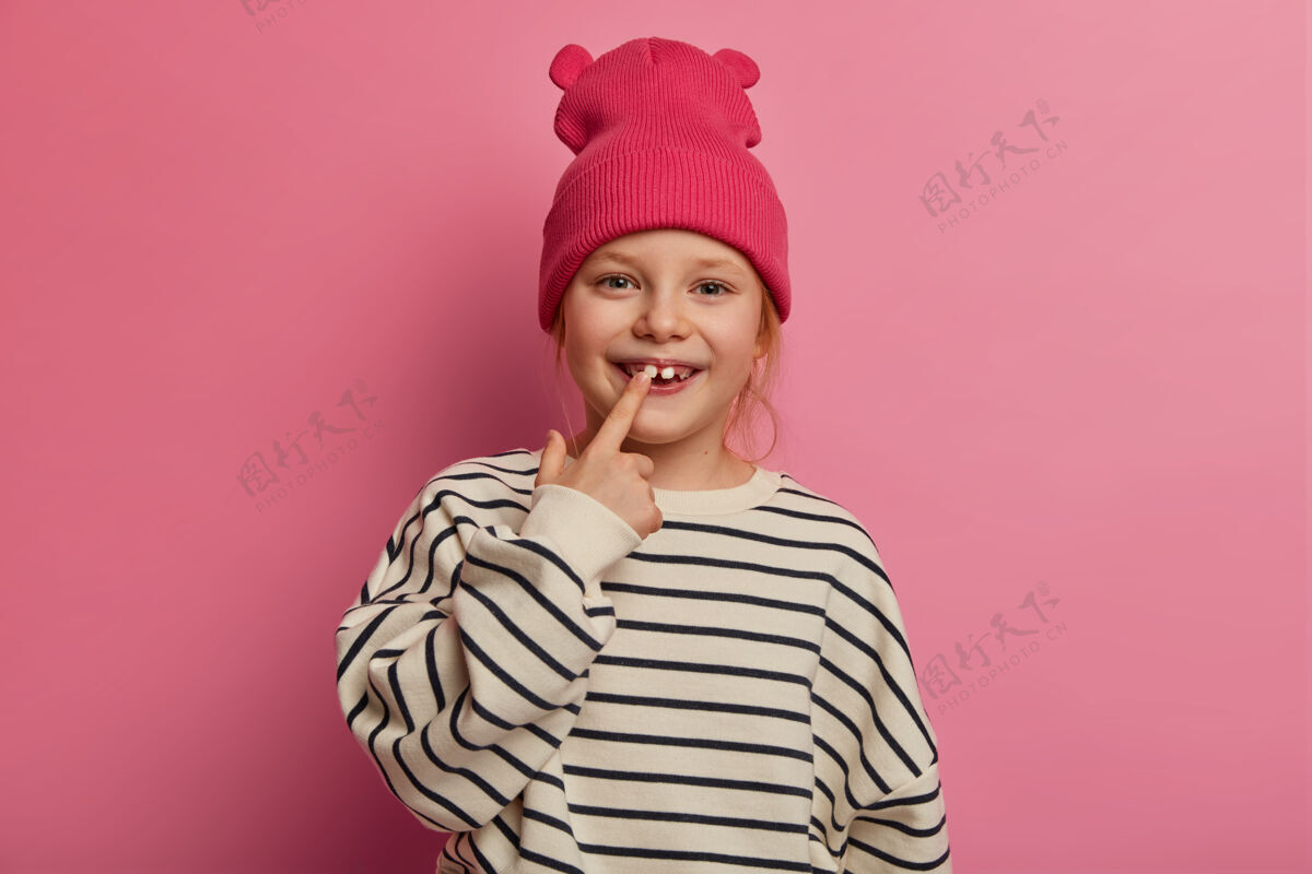 高兴快乐有趣的小女孩指着自己的牙齿 关心口腔卫生 穿着时髦的衣服 有健康的皮肤 在操场上向朋友炫耀自己的成人牙齿 隔离在粉色粉彩墙上可爱时尚罗西