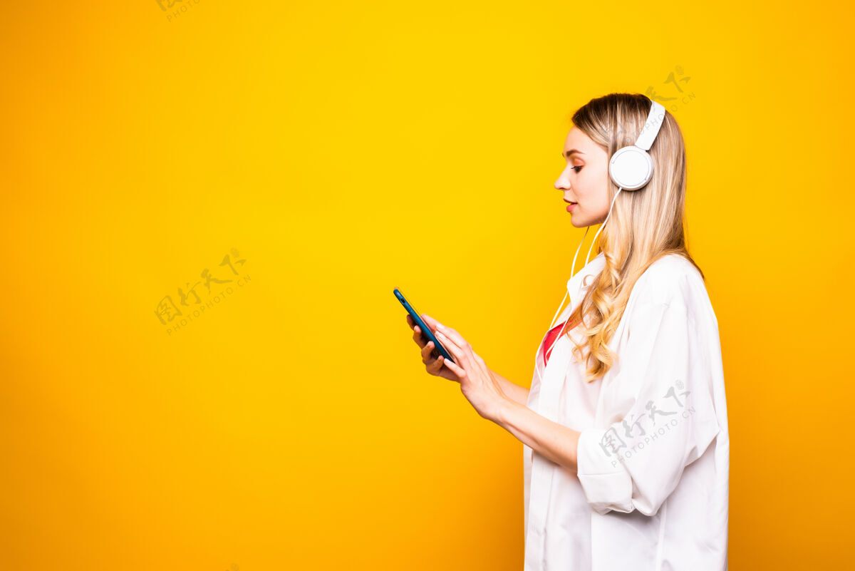肖像一个戴着耳机和手机隔着黄墙听音乐的快乐年轻女子的画像手手机电话