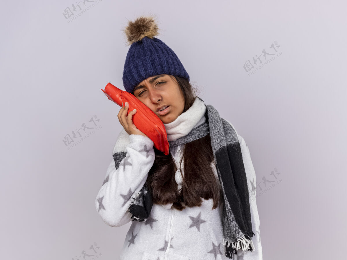 围巾悲伤的生病的年轻女孩戴着冬天的帽子戴着围巾把热水袋放在脸颊上隔离在白色的背景上脸颊女孩包