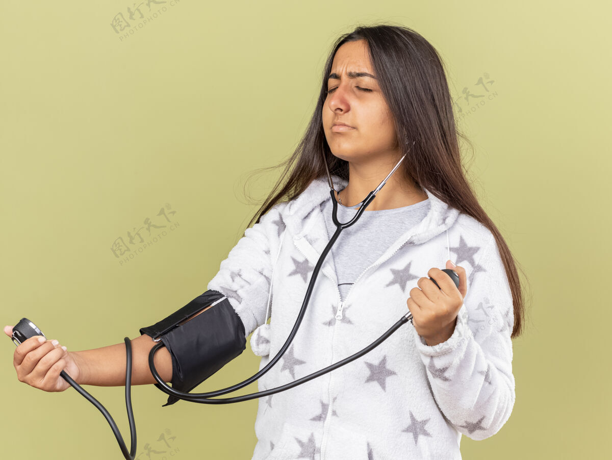 橄榄年轻的病女孩闭着眼睛用橄榄绿背景上隔离的血压计测量自己的血压自己年轻压力