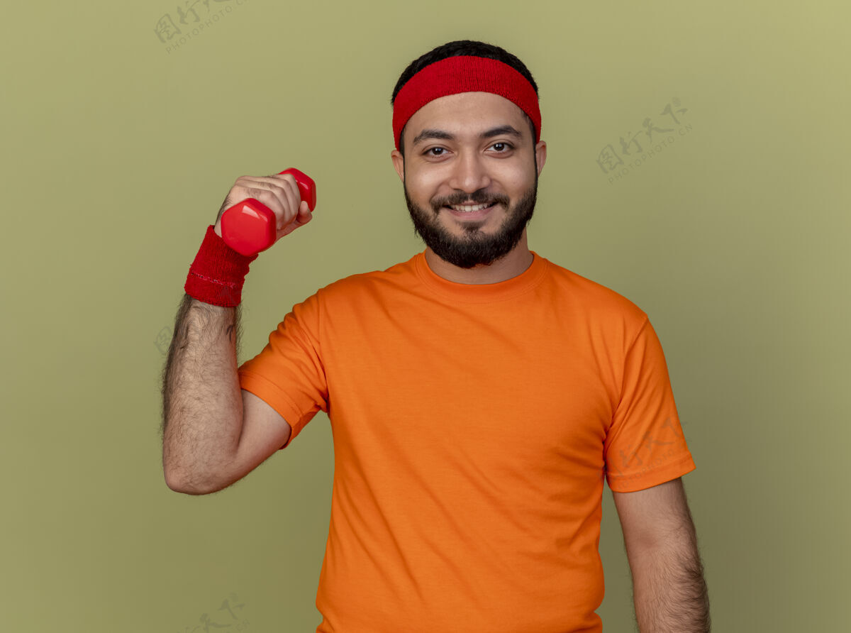 锻炼微笑的年轻运动男子戴着头带和腕带锻炼哑铃孤立在橄榄绿的背景橄榄腕带头带