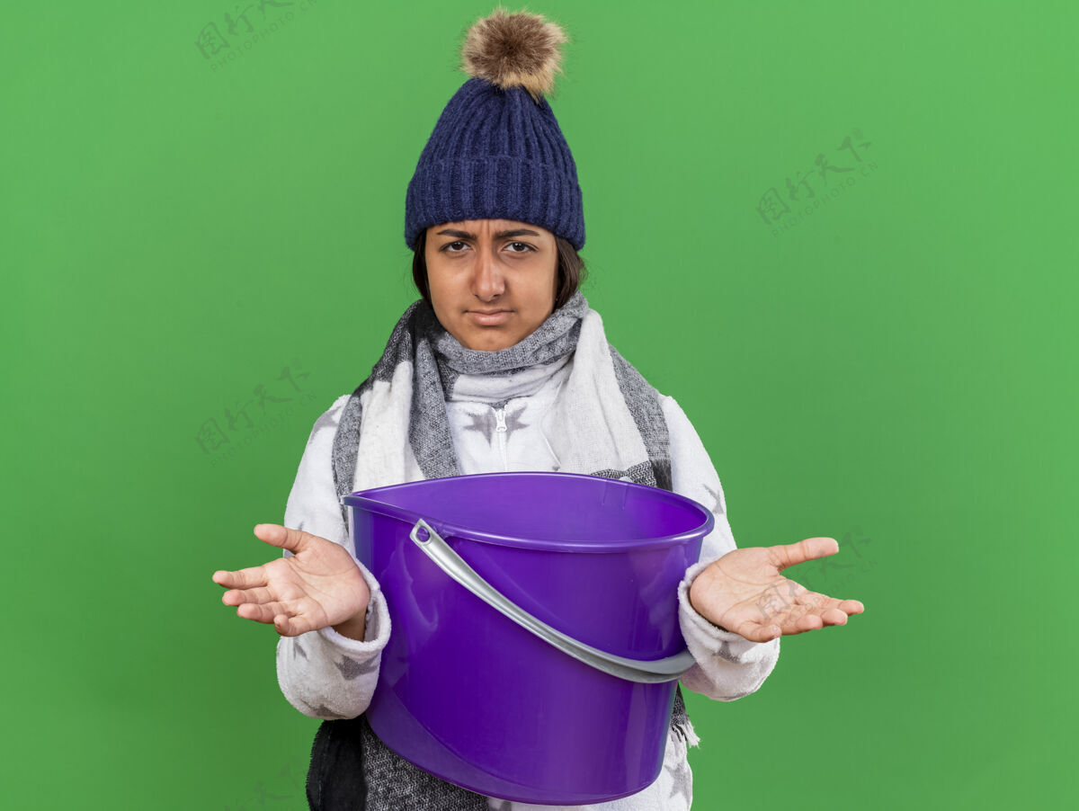 不愉快戴着冬天的帽子戴着围巾拿着塑料桶摊开双手隔离在绿色背景下的年轻病女孩帽子持有疾病