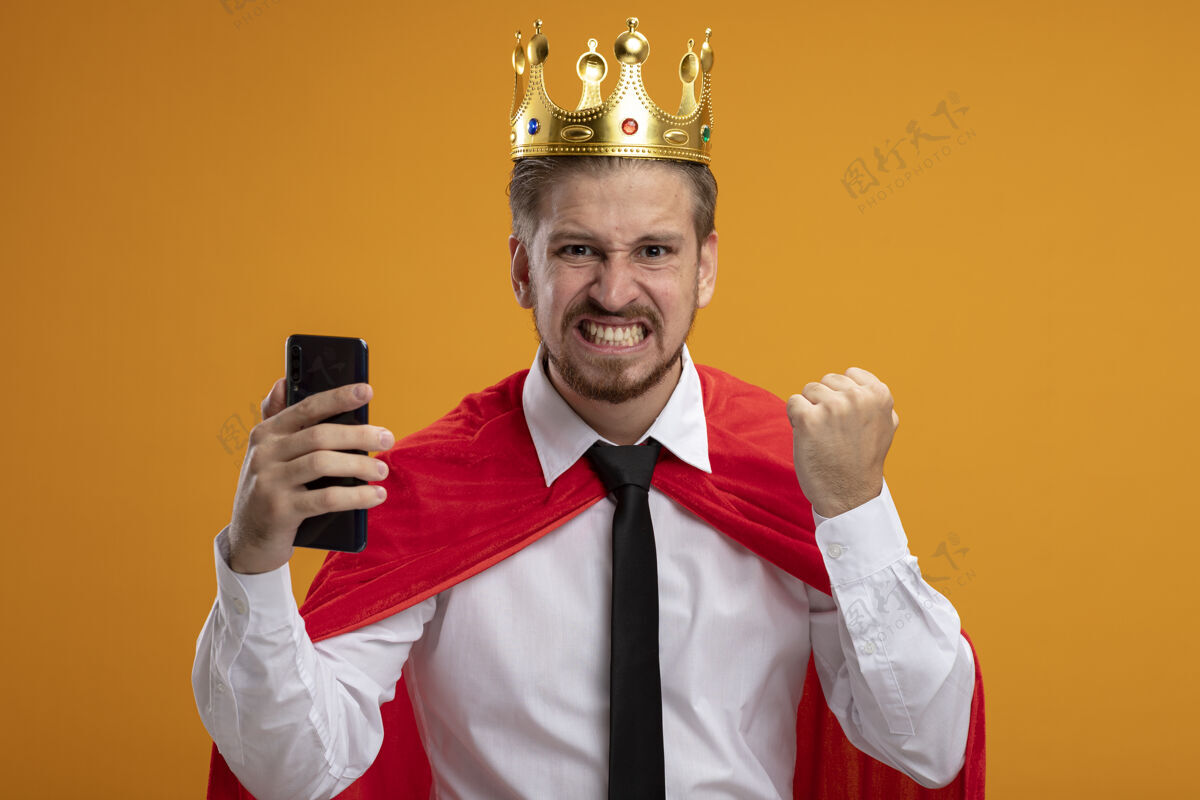 快乐快乐的年轻超级英雄戴着领带 戴着王冠 拿着电话 在橙色背景上显示“是”的手势领带抱着皇冠