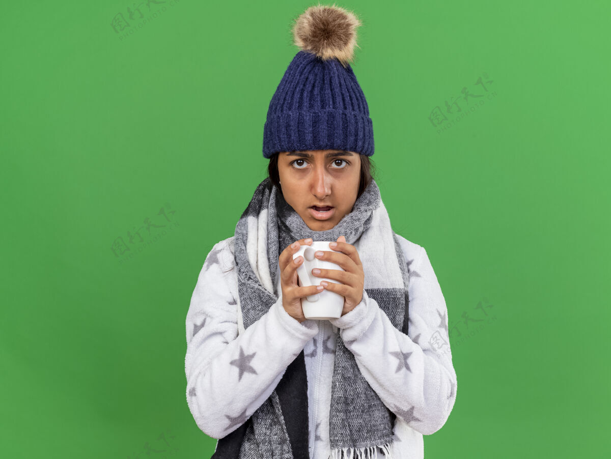 帽子惊讶的年轻生病的女孩戴着冬天的帽子 戴着围巾 端着一杯茶 隔离在绿色的背景上惊喜女孩抱着